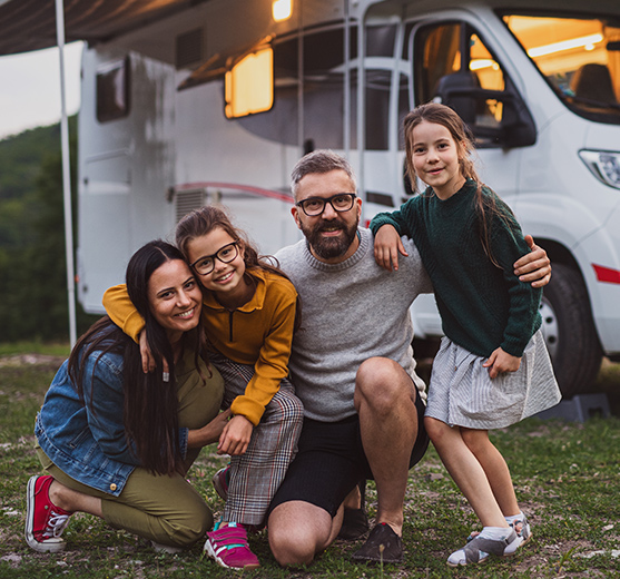 Middle-aged parents with two young girls are posting in front of a camper around dusk.