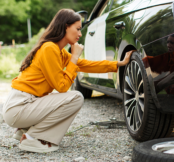 Stressed woman looking at flat tire and thinking how to change wheel of car, sitting on roadside outdoors
