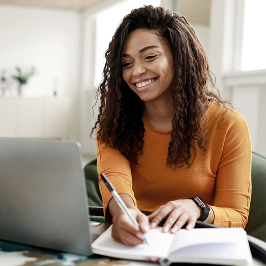 Young woman sitting at desk working on laptop taking notes in notebook