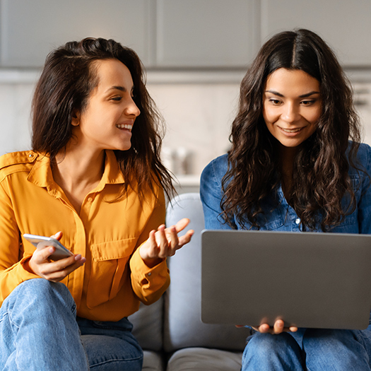 Two smiling women sitting comfortably on a couch engaging with a laptop