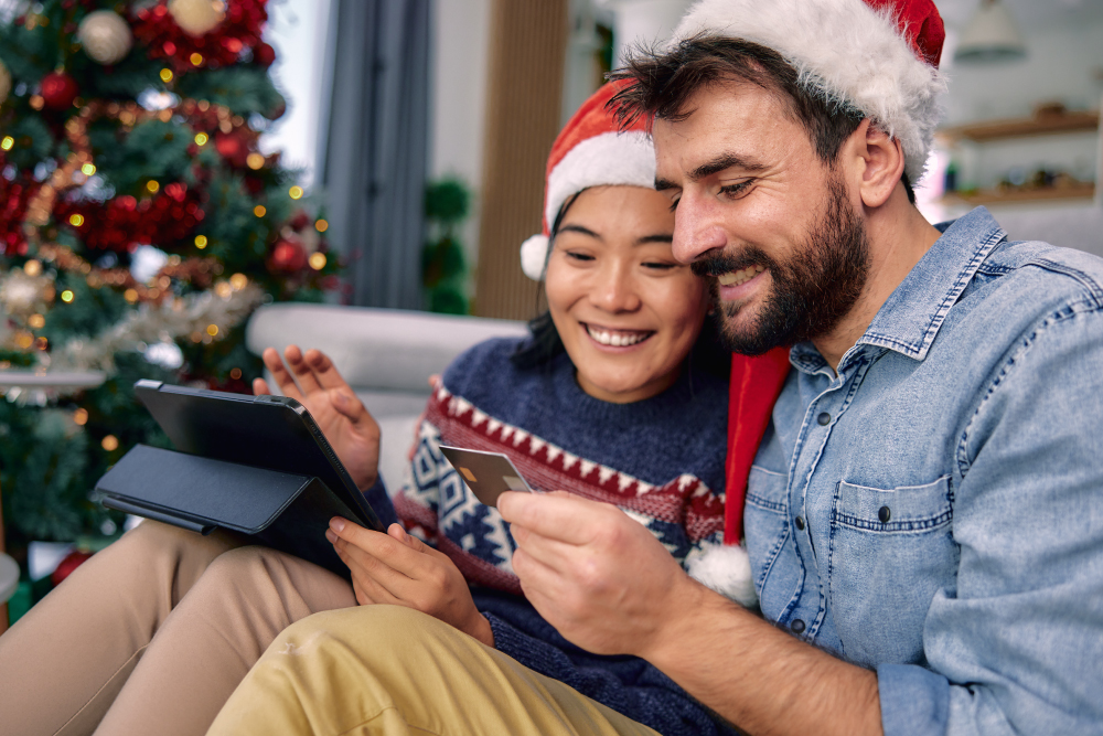 A couple dressed in holiday attire looking at their credit card while buying something online at the same time.